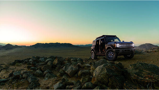 A Ford Bronco with doors removed driving in the desert with mountains in the background at sundown.